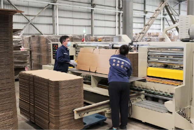 Corrugated cardboard manufacturing process at SC PACK BOX facility with workers operating packaging machinery.
